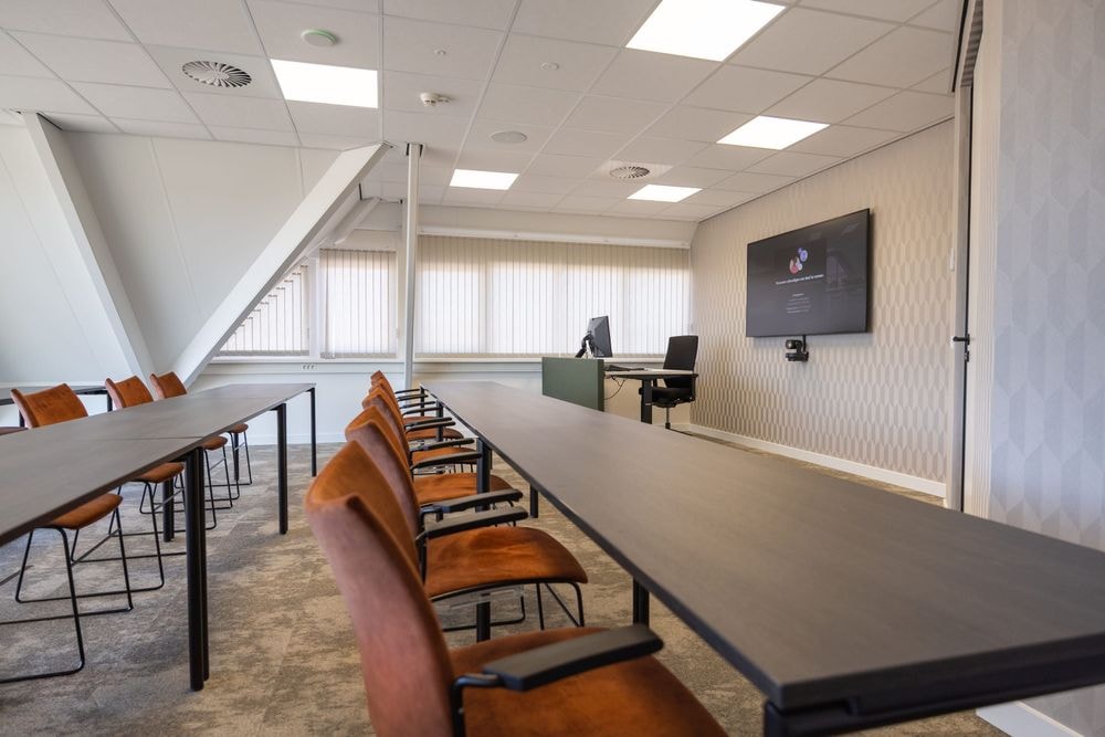 Harry Frens kamer in cursusopstelling voor 18 personen met tafels, stoelen, 75-inch scherm en ramen met daglicht — Mudde Kok FlexCentre
